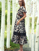 A woman in a chic leopard print dress stands amidst trees, blending nature with her stylish attire.