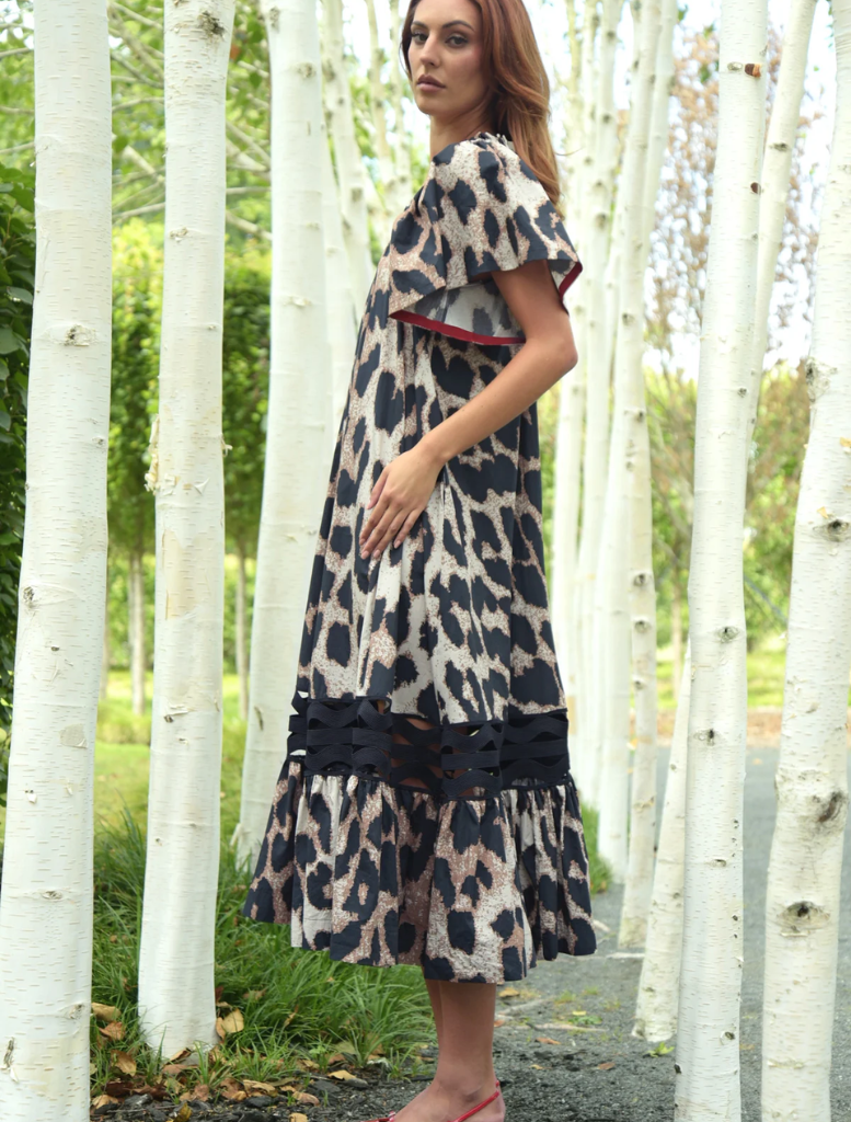 A woman in a chic leopard print dress stands amidst trees, blending nature with her stylish attire.