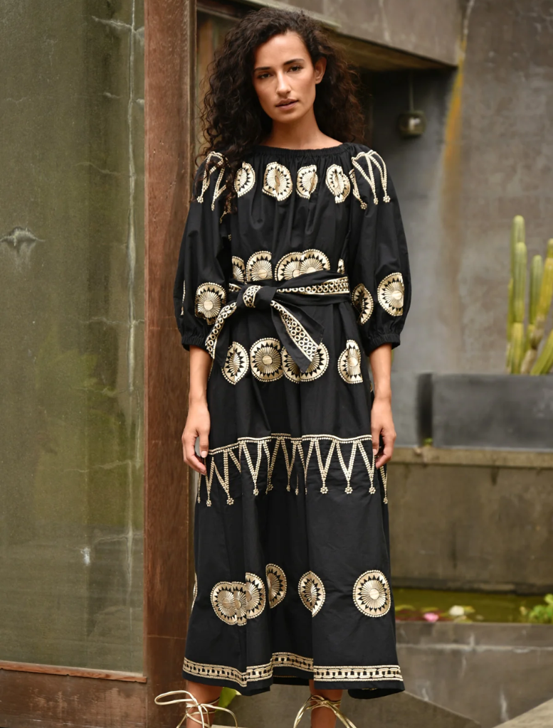 A woman in a sleek black dress leans against a wall, highlighting her poised and sophisticated appearance.