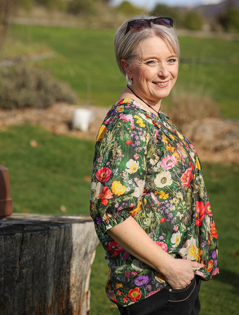 The model is dressed in a floral blouse and wide-leg pants, featuring the Looking Glass Top in Ivy Multi color scheme.