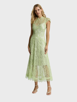  A woman models the Lorena Midi Dress in citrus, standing in a garden filled with vibrant flowers and greenery.
