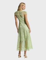 A woman in a lace dress named Lorena Midi Dress - Citrus stands in a blooming garden, exuding elegance and style.