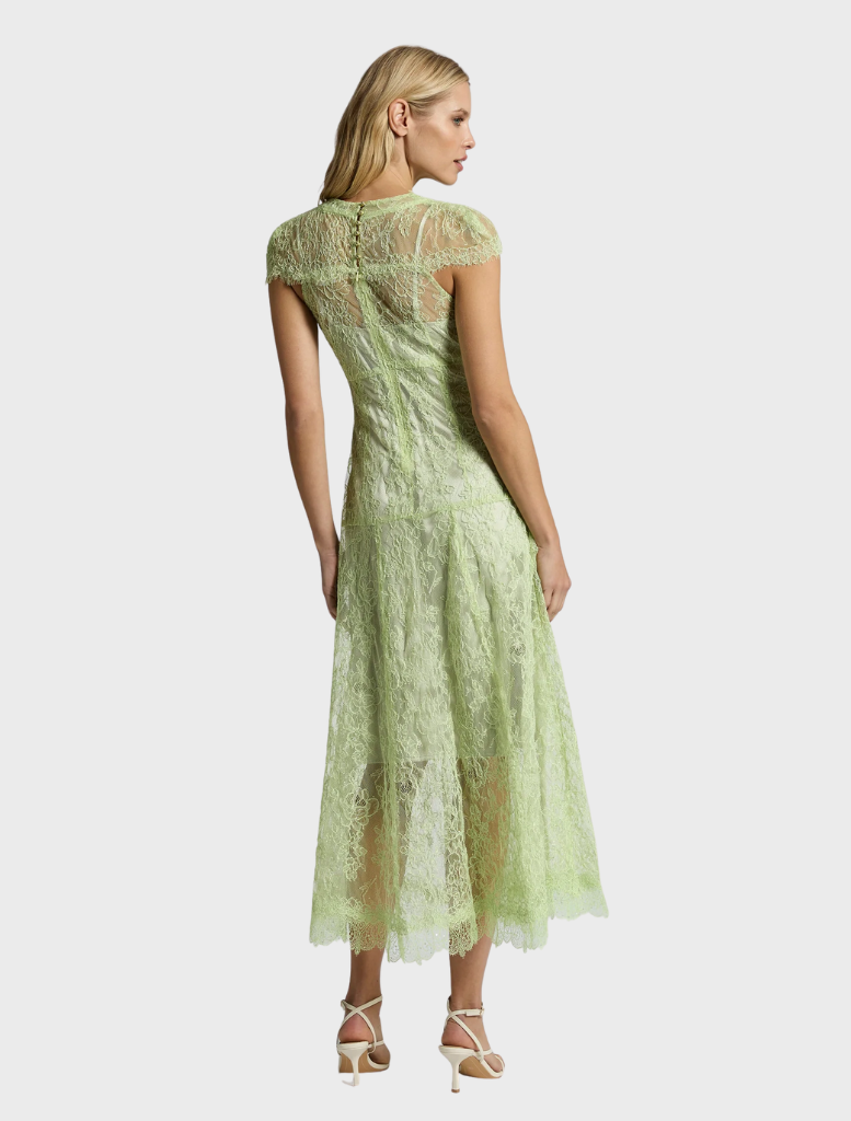 A woman in a lace dress named Lorena Midi Dress - Citrus stands in a blooming garden, exuding elegance and style.