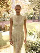 A woman in a citrus-colored lace dress stands gracefully in a vibrant garden, surrounded by blooming flowers.