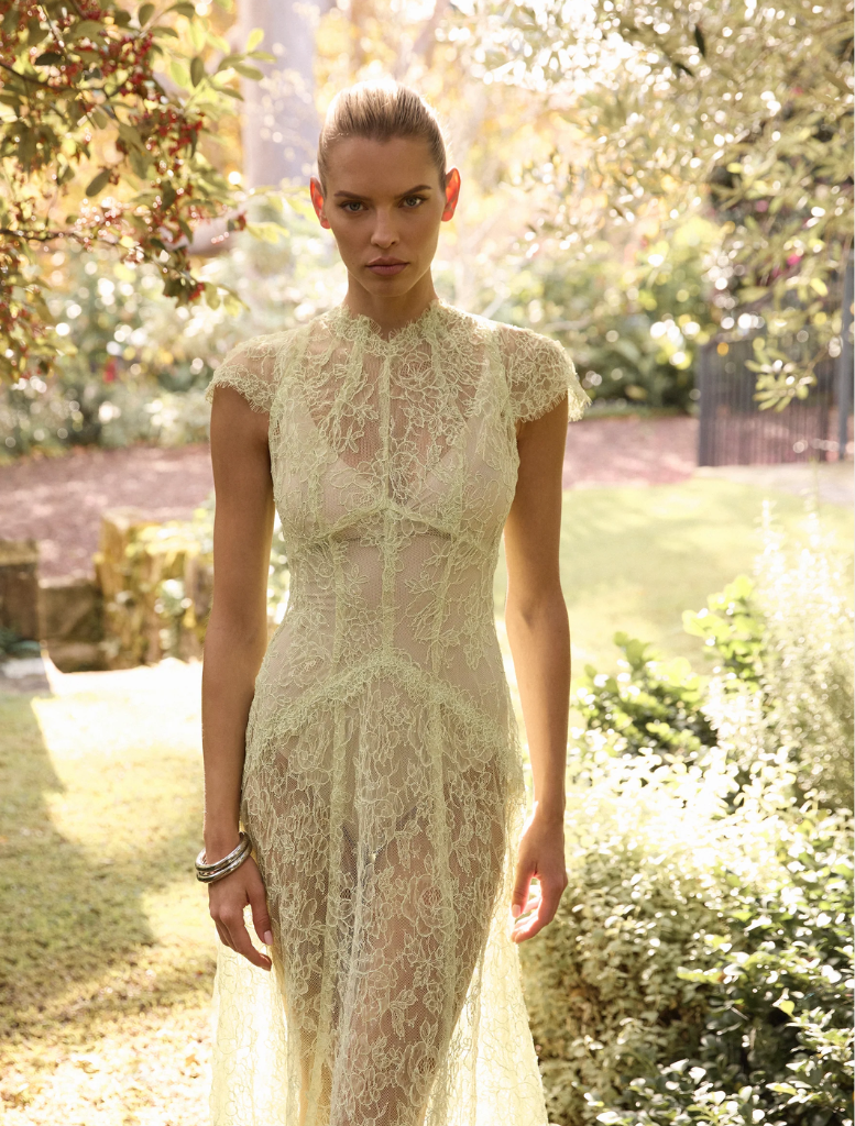 A woman in a citrus-colored lace dress stands gracefully in a vibrant garden, surrounded by blooming flowers.