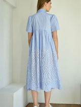 A woman dressed in a blue gown with intricate white lace embellishments smiles softly.