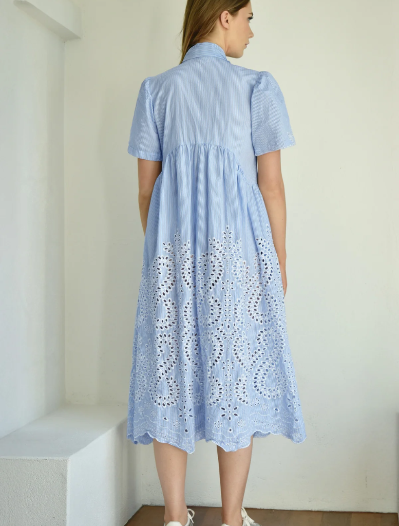 A woman dressed in a blue gown with intricate white lace embellishments smiles softly.