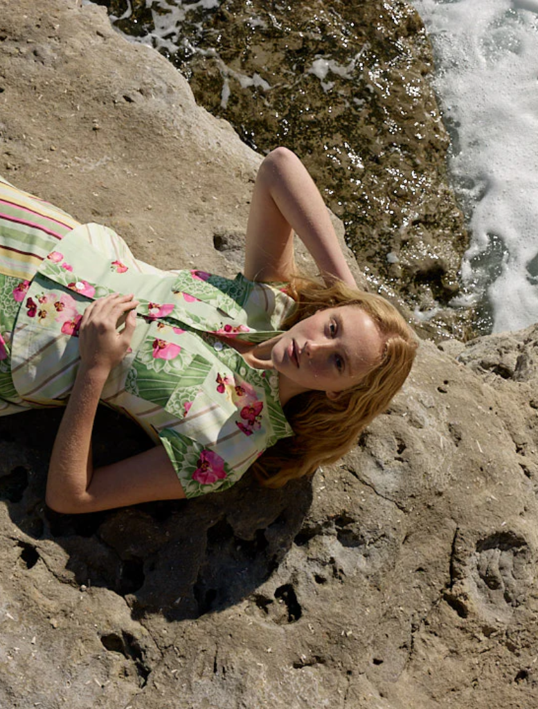A woman in a floral dress, Tilla Denom Top - Meadow Multi, stands on a rocky beach, surrounded by natural coastal scenery.