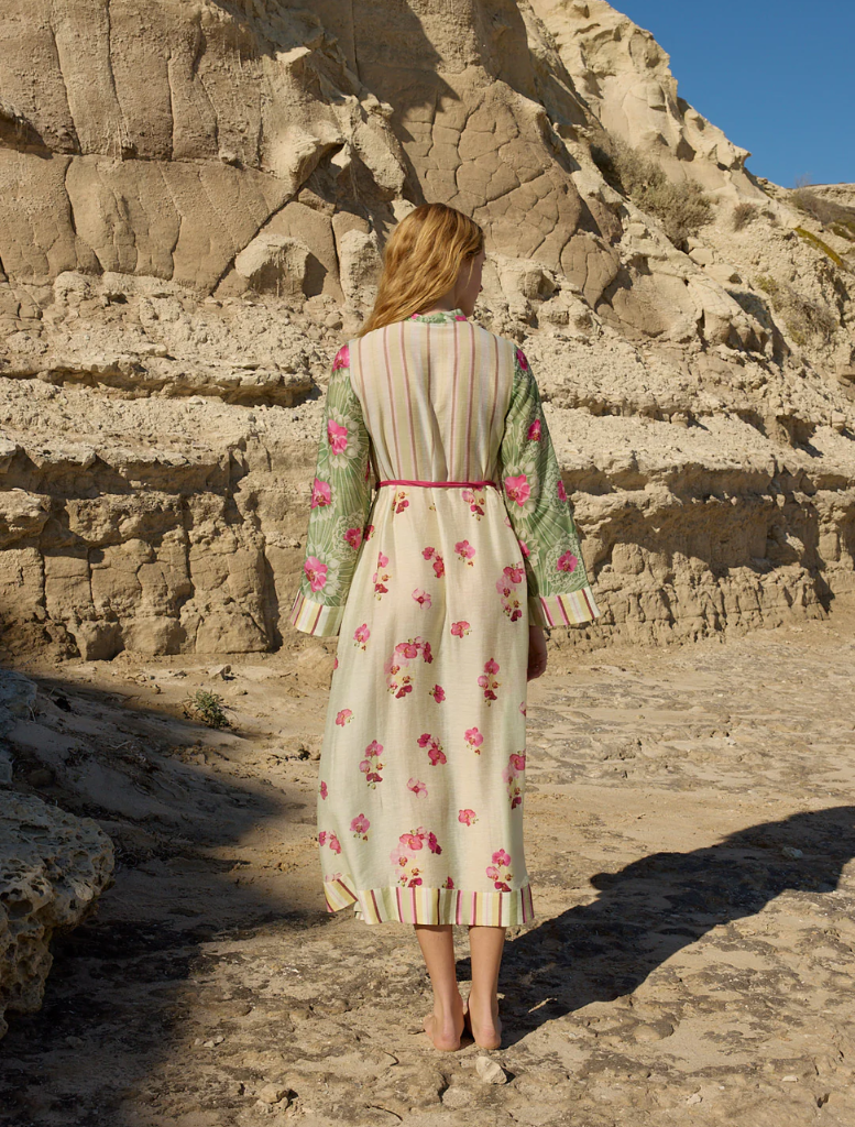 A woman in a floral dress, the Neottia Dress - Meadow Phalae, strolls along the sandy beach under a clear sky.