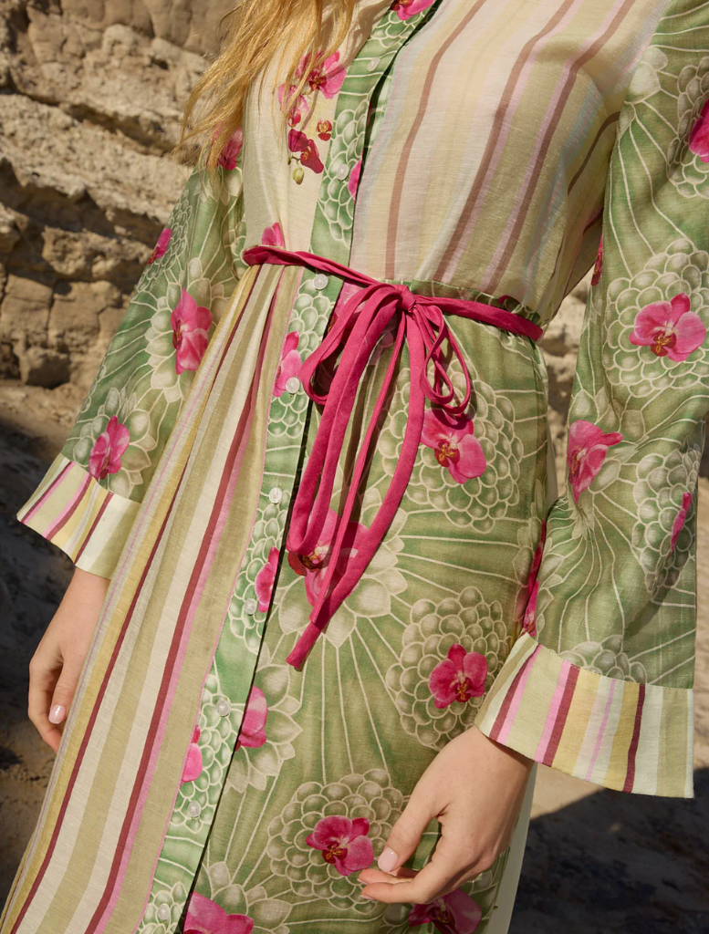 A woman wearing the Neottia Dress - Meadow Phalae stands on a beach, surrounded by sand and waves under a clear sky.
