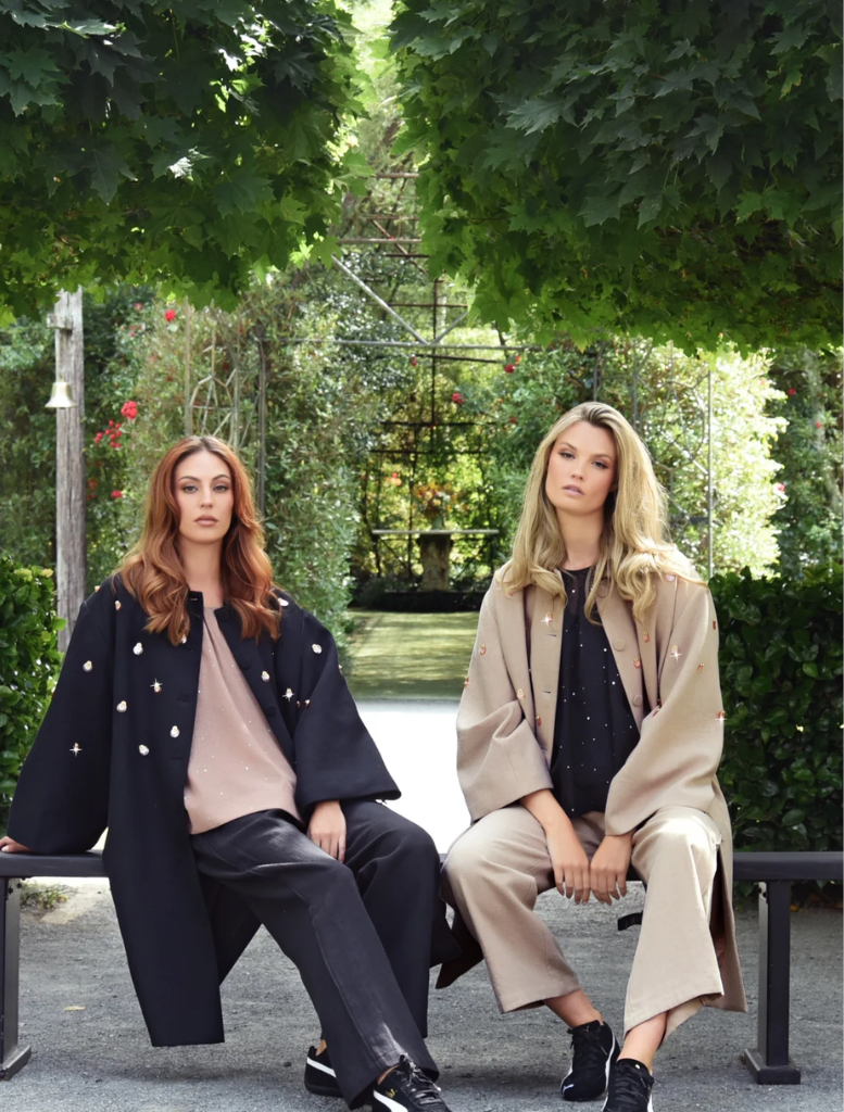 Two women wearing black trousers sit on a bench in a lush garden, enjoying a peaceful moment together.