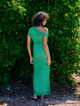 A woman dressed in green stands elegantly before a backdrop of leafy bushes.