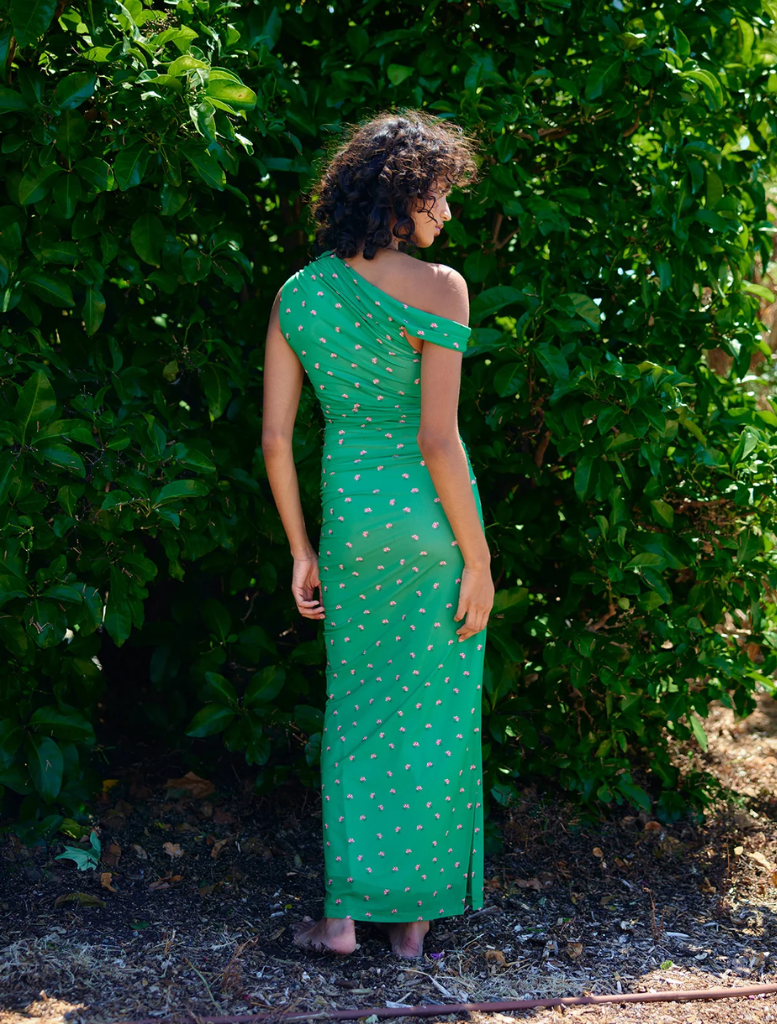 A woman dressed in green stands elegantly before a backdrop of leafy bushes.