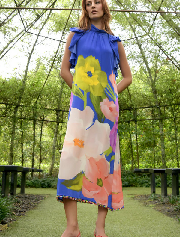 A woman in a cobalt floral dress and pink sandals stands gracefully in a vibrant garden setting.