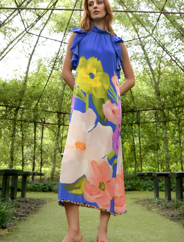 A woman in a cobalt floral dress and pink sandals stands gracefully in a vibrant garden setting.