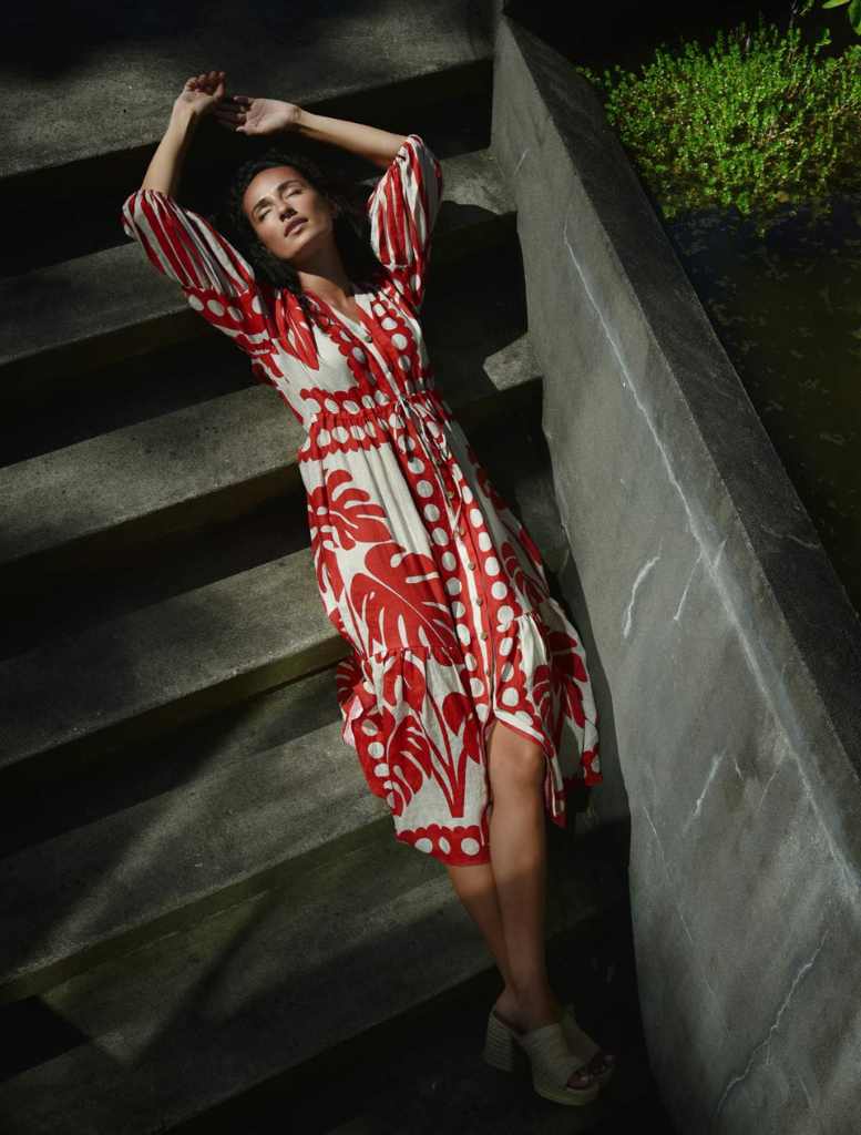 A woman wearing a red and white dress poses in front of a plain wall, her hands resting at her sides.