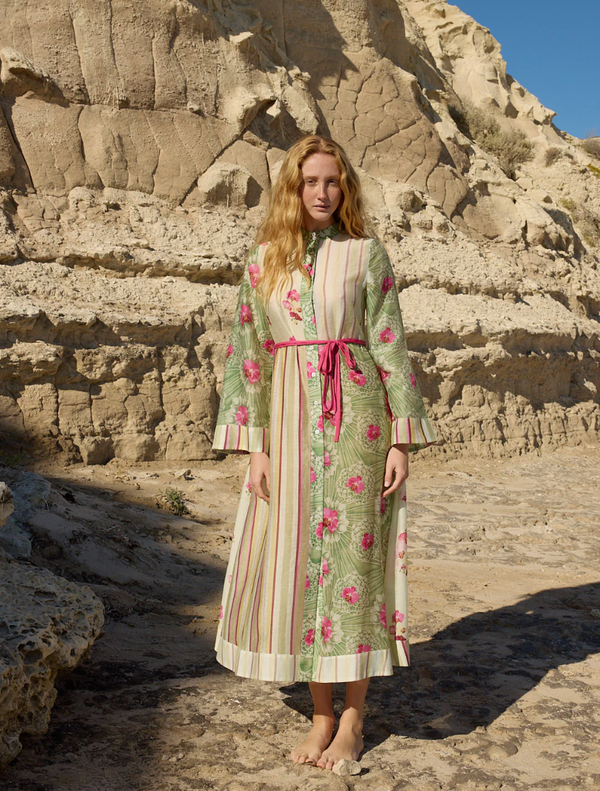 A woman in a floral dress stands on a beach, showcasing the Neottia Dress - Meadow Phalae against a serene ocean backdrop.
