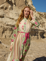  On a beach, a woman in a floral Neottia Dress - Meadow Phalae poses, with the ocean and horizon visible in the background.