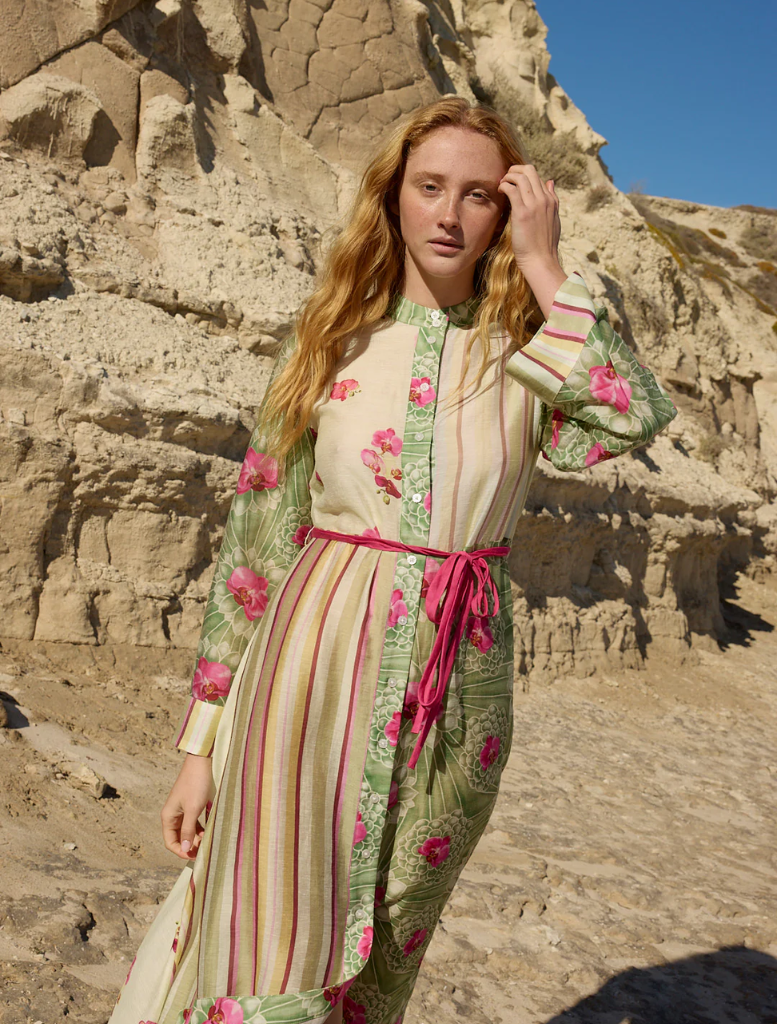  On a beach, a woman in a floral Neottia Dress - Meadow Phalae poses, with the ocean and horizon visible in the background.