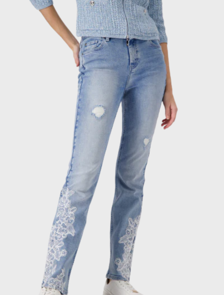 A woman in jeans featuring intricate lace detailing on the pockets and hem, standing confidently with a neutral background.