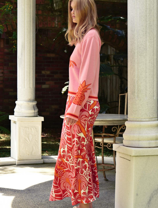 A woman dressed in a pink sweater paired with a red skirt poses, highlighting her colorful fashion choice.