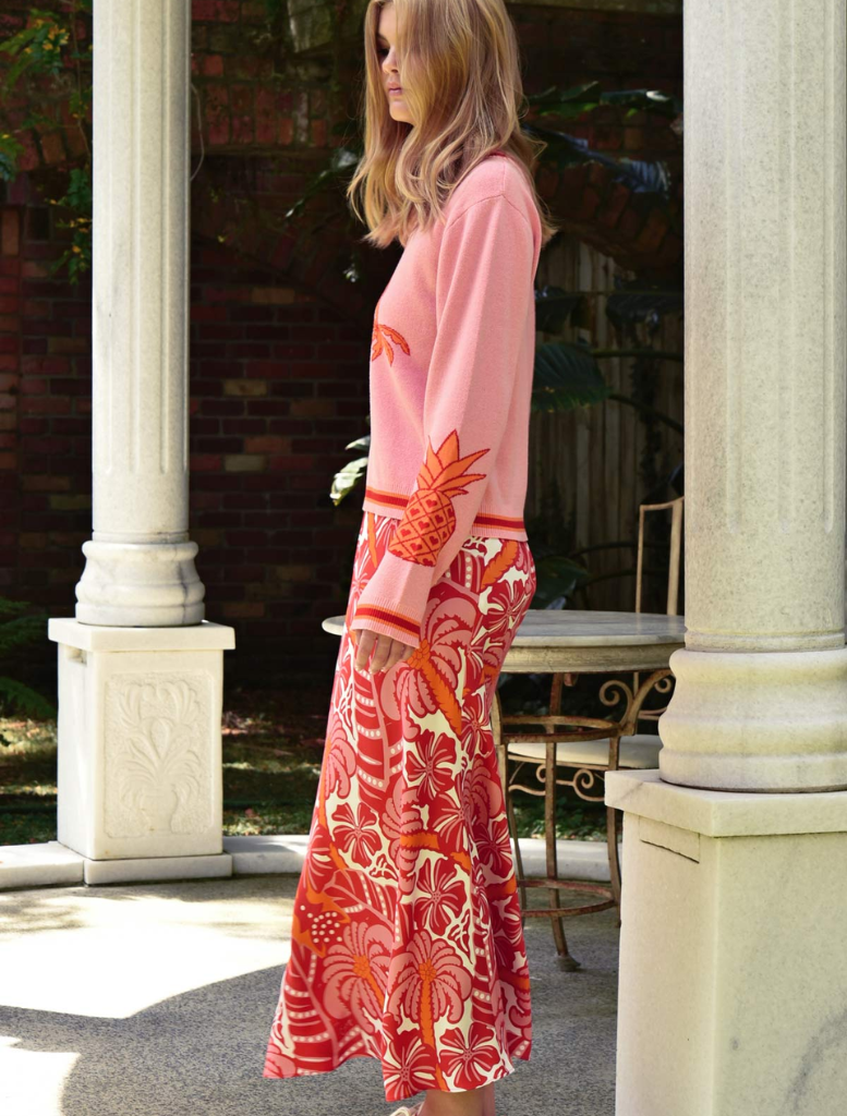 A woman dressed in a pink sweater paired with a red skirt poses, highlighting her colorful fashion choice.
