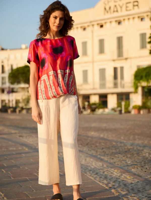 A woman in a vibrant top and white pants stands confidently, showcasing her colorful outfit against a neutral background.
