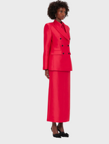 A woman wearing a red suit over a black shirt, standing confidently with a neutral expression.