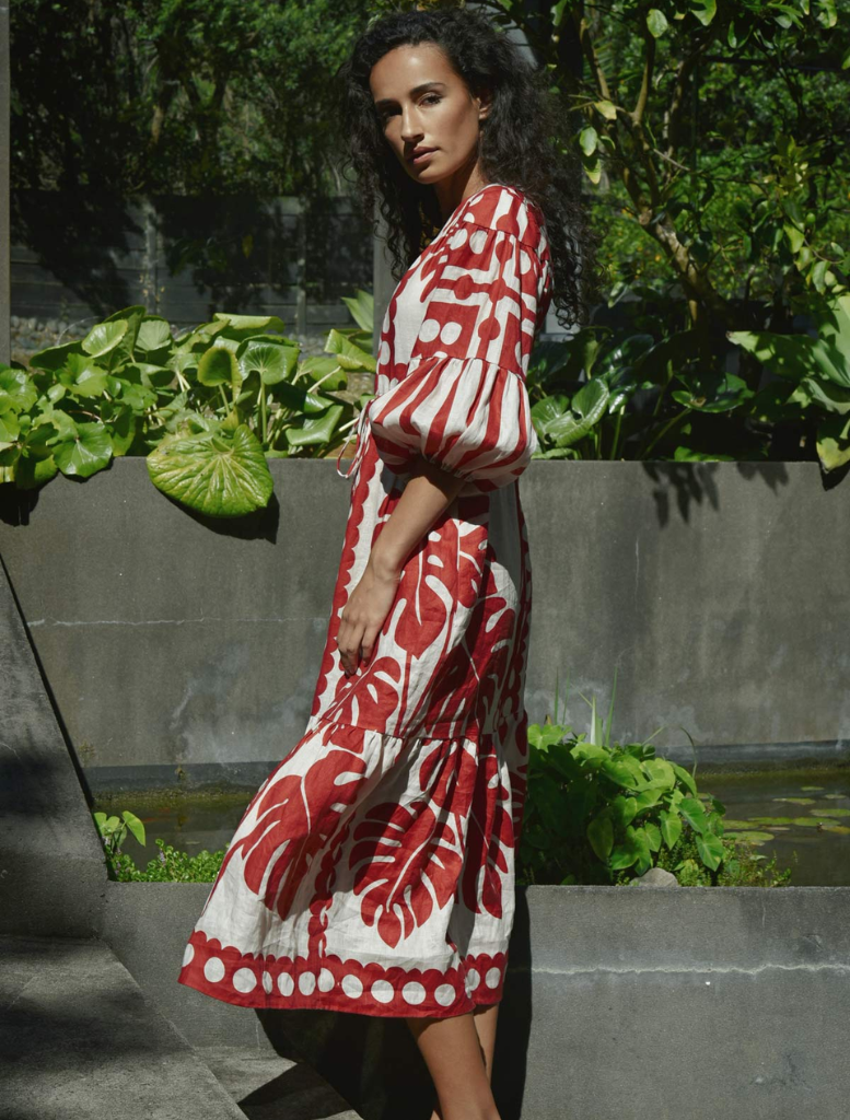 A woman wearing a red and white dress poses in front of a plain wall, her hands resting at her sides.