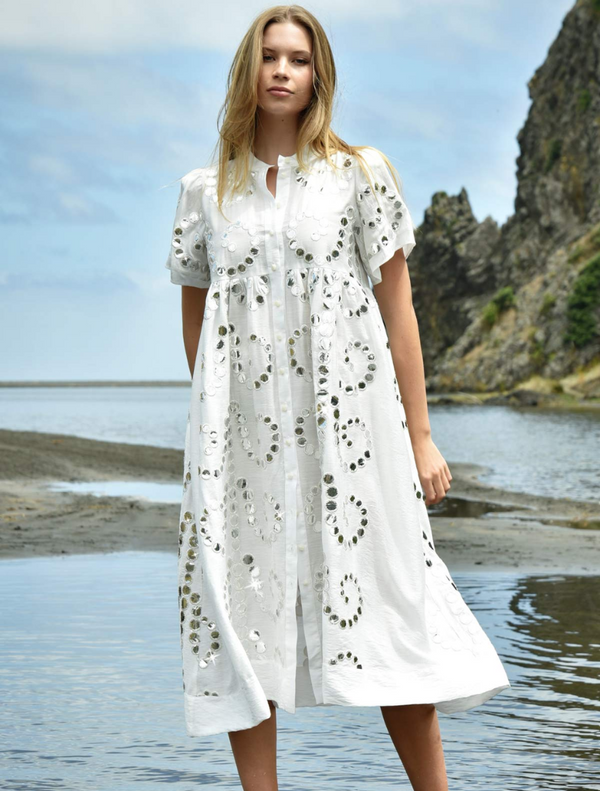 A woman in a flowing white dress stands gracefully on the beach, with waves gently lapping at her feet.