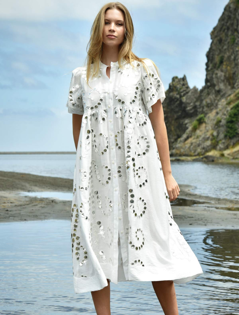 A woman in a flowing white dress stands gracefully on the beach, with waves gently lapping at her feet.