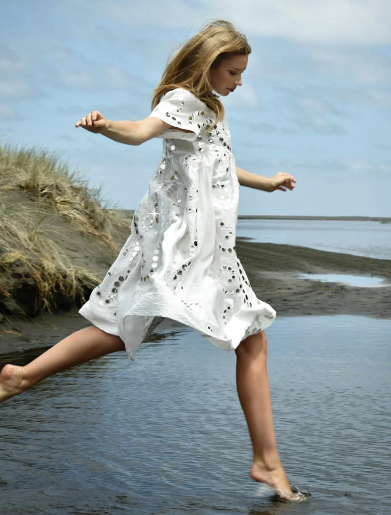 A woman in a white dress stands on the beach, surrounded by sand and water, enjoying the serene coastal view.