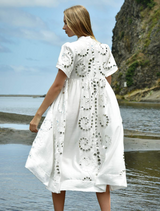 A woman in a white dress stands on the beach, her hair blowing in the wind as she looks out at the sea.