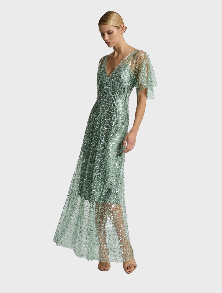 A woman stands in a garden wearing a sage Tilda gown, with greenery and flowers creating a serene and picturesque backdrop.