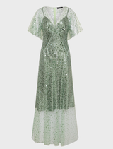 A woman wearing a long sage Tilda gown stands in a blooming garden, showcasing the beauty of nature around her.
