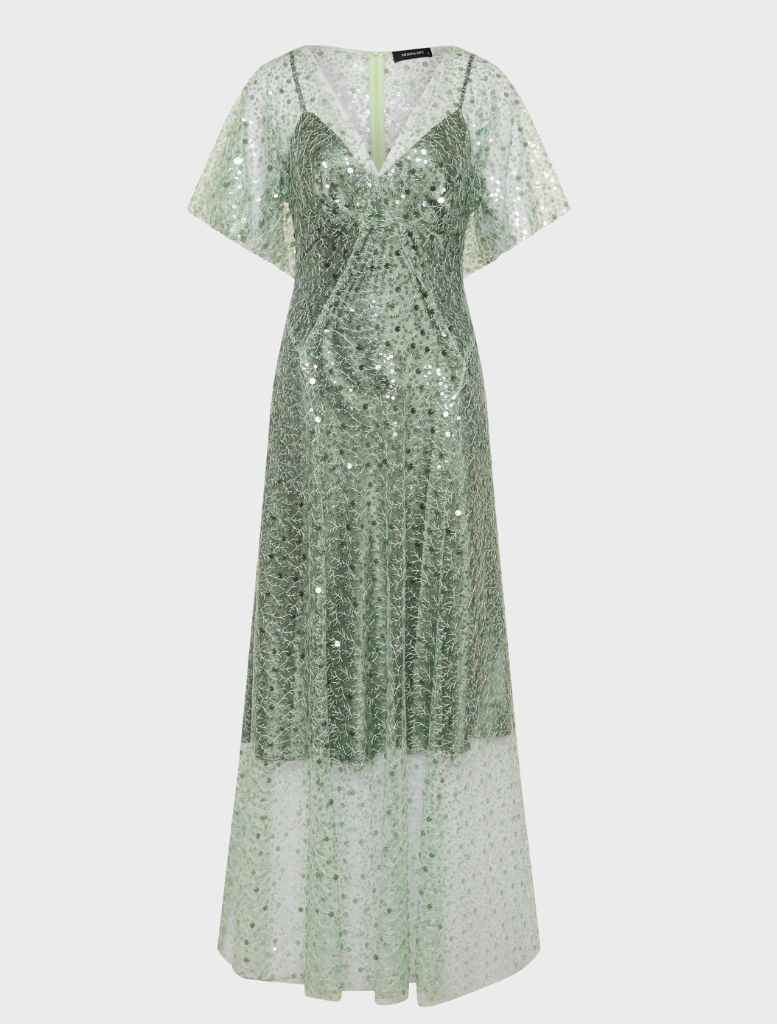 A woman wearing a long sage Tilda gown stands in a blooming garden, showcasing the beauty of nature around her.
