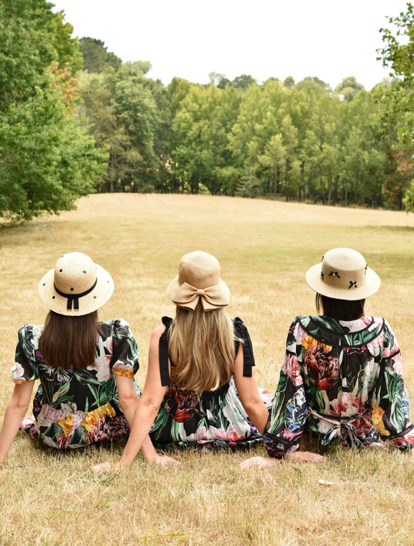 Three women in floral dresses and straw hats sitting on the grass, enjoying a sunny day outdoors.