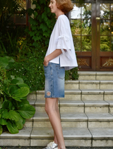 A woman dressed in a white shirt and denim shorts poses in a sunny outdoor setting.