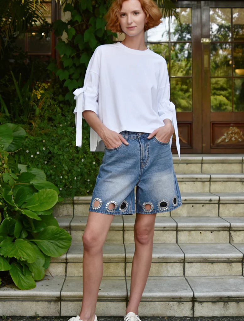 A woman wearing a white shirt and denim shorts stands casually outdoors.