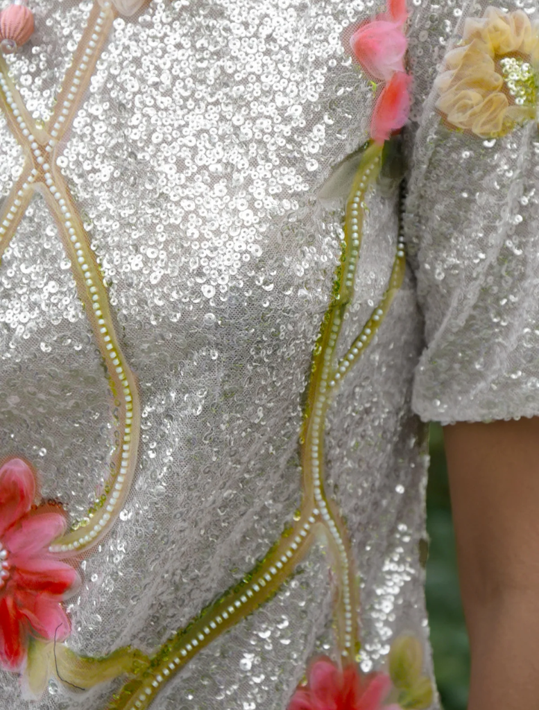 A woman in a floral dress of silver and pink leans against a tree, capturing a tranquil moment in a natural environment.
