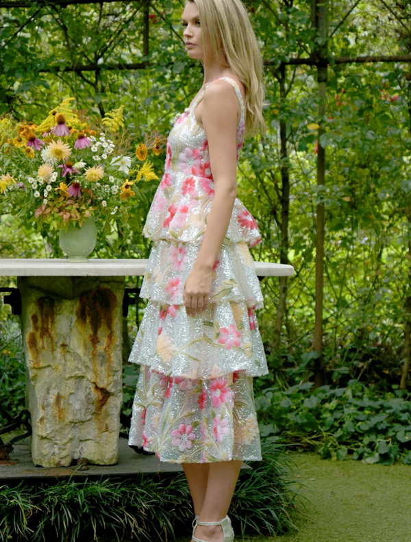 A woman wearing a floral dress poses in front of a lush garden, surrounded by colorful flowers and greenery.