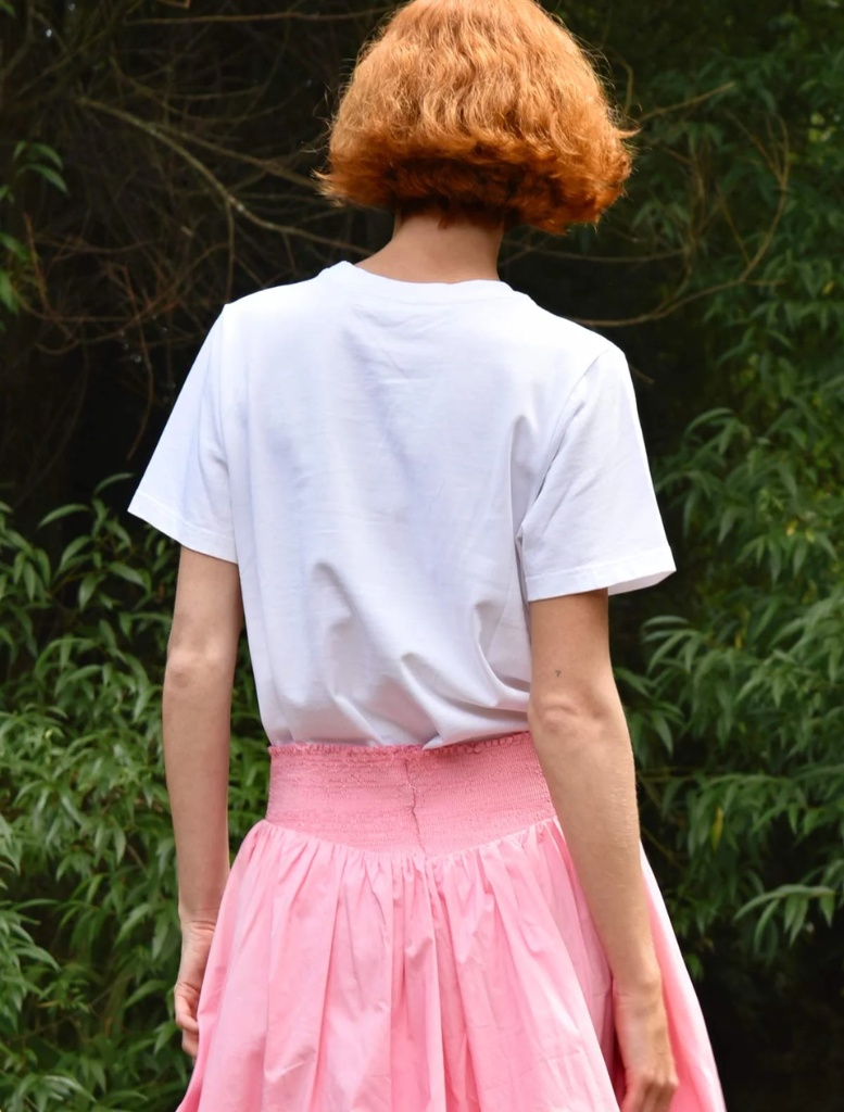 A woman dressed in a pink skirt and a white t-shirt adorned with a bow stands against a vibrant background.