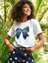 A woman in a white t-shirt adorned with a bow, smiling and standing against a neutral background.