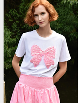 A woman in a pink skirt and a white t-shirt featuring a bow stands smiling outdoors.