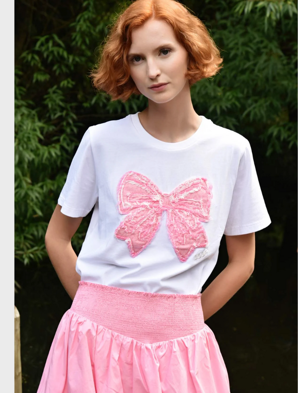 A woman in a pink skirt and a white t-shirt featuring a bow stands smiling outdoors.