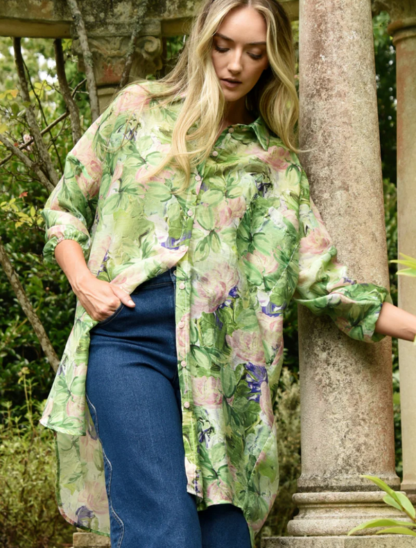 A woman in a green floral shirt and jeans leans against a pillar, showcasing a casual and stylish look.
