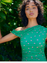 A woman in a flowing green dress stands amidst dense green bushes.