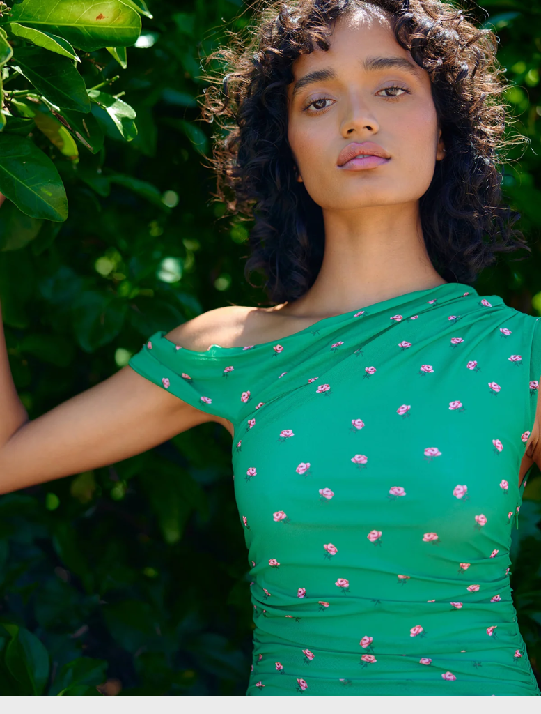A woman in a flowing green dress stands amidst dense green bushes.