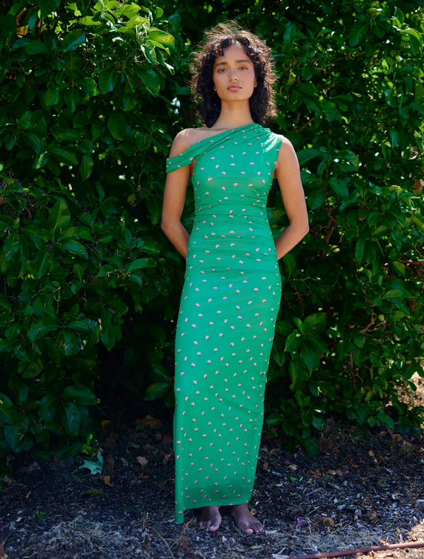 A woman in a green dress stands gracefully in front of lush green bushes.
