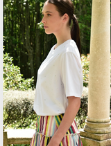 A woman in a white Sparkle Magic T-shirt paired with a vibrant multicolored striped skirt poses cheerfully outdoors.